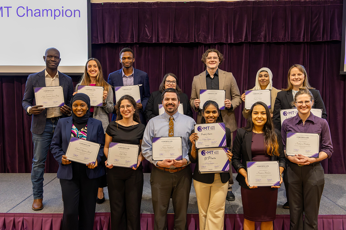About a dozen graduate students on a platform hold certificates at a research presentation competition in a ballroom.