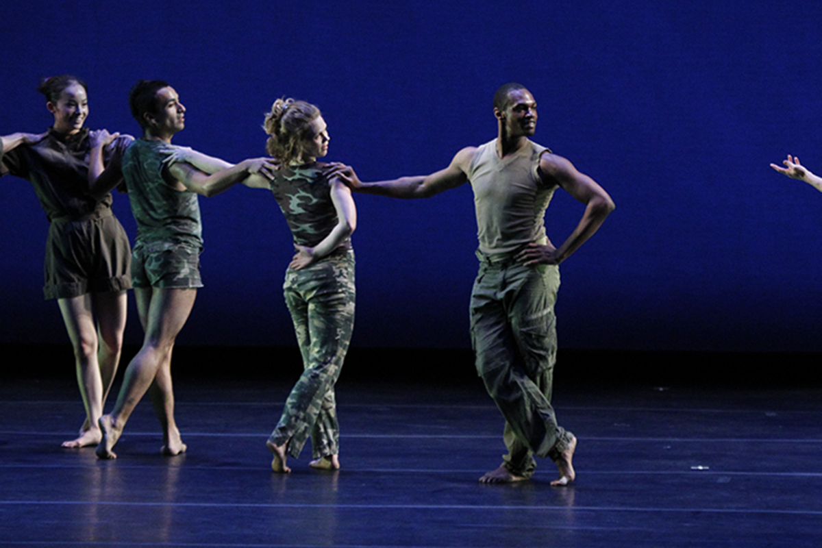 A group of dancers on a darkly lit stage with a blue background strike a dramatic pose in a line.