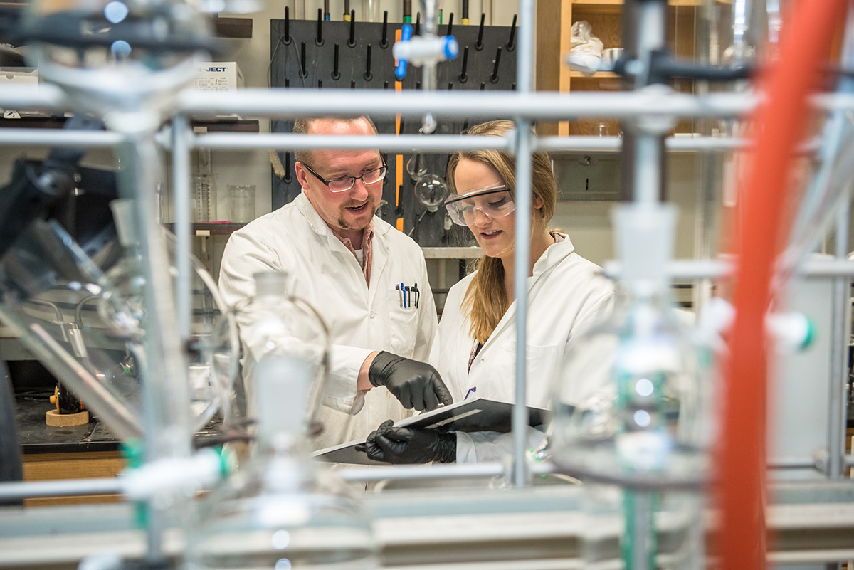 A man wearing a white lab coat and a young woman wearing a white lab coat stand in a lab partially obscured by piping and tubes. The man is pointing to a document the woman is holding.