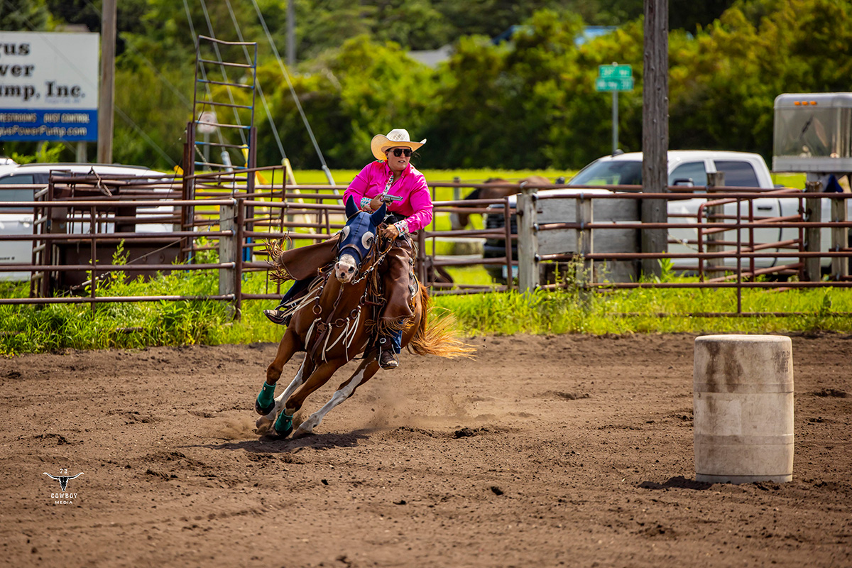 A woman in a pink western shirt rides a brown horse and competes in a mounted shooting competition. 