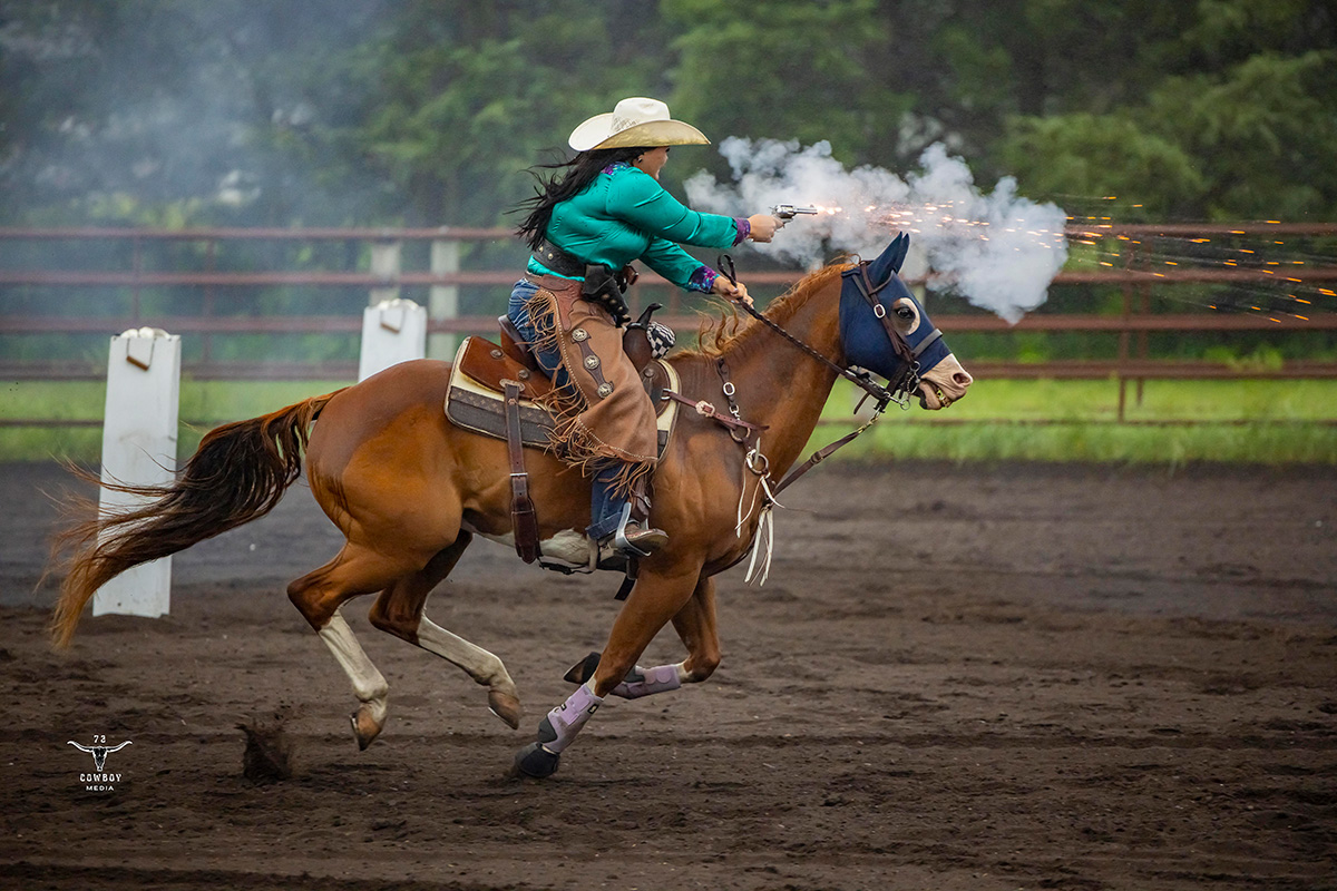 A college student competes while riding a brown horse and shooting a rifle at a balloon. 