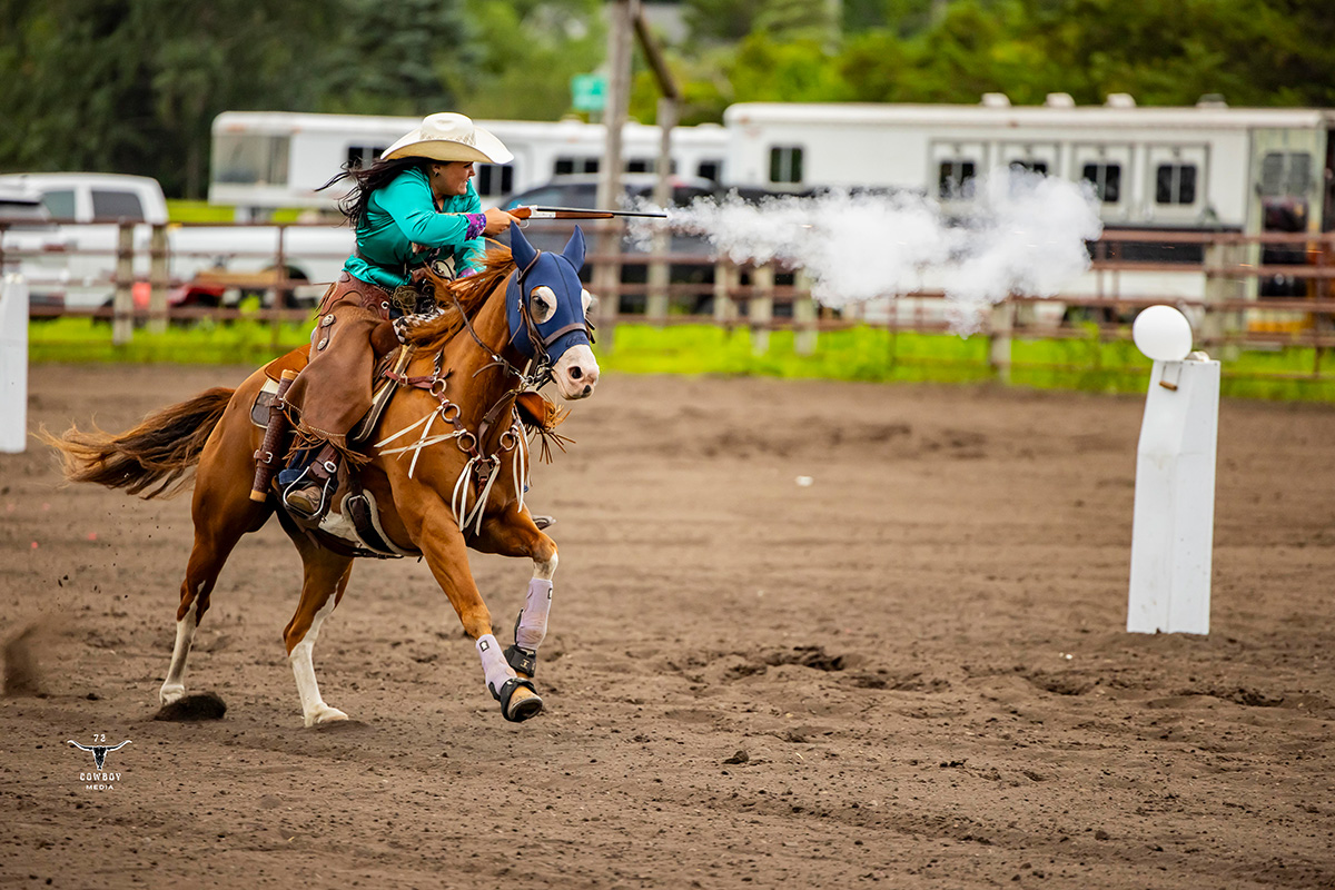 A college student competes in mounted shooting while riding a brown horse in an arena. 
