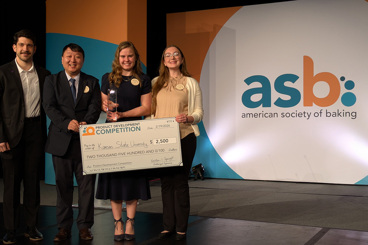 Four college students in business attire stand together on a stage holding a large check and a crystal award. In the background, an orange and teal banner with the American Society of Baking conference initials is displayed. 