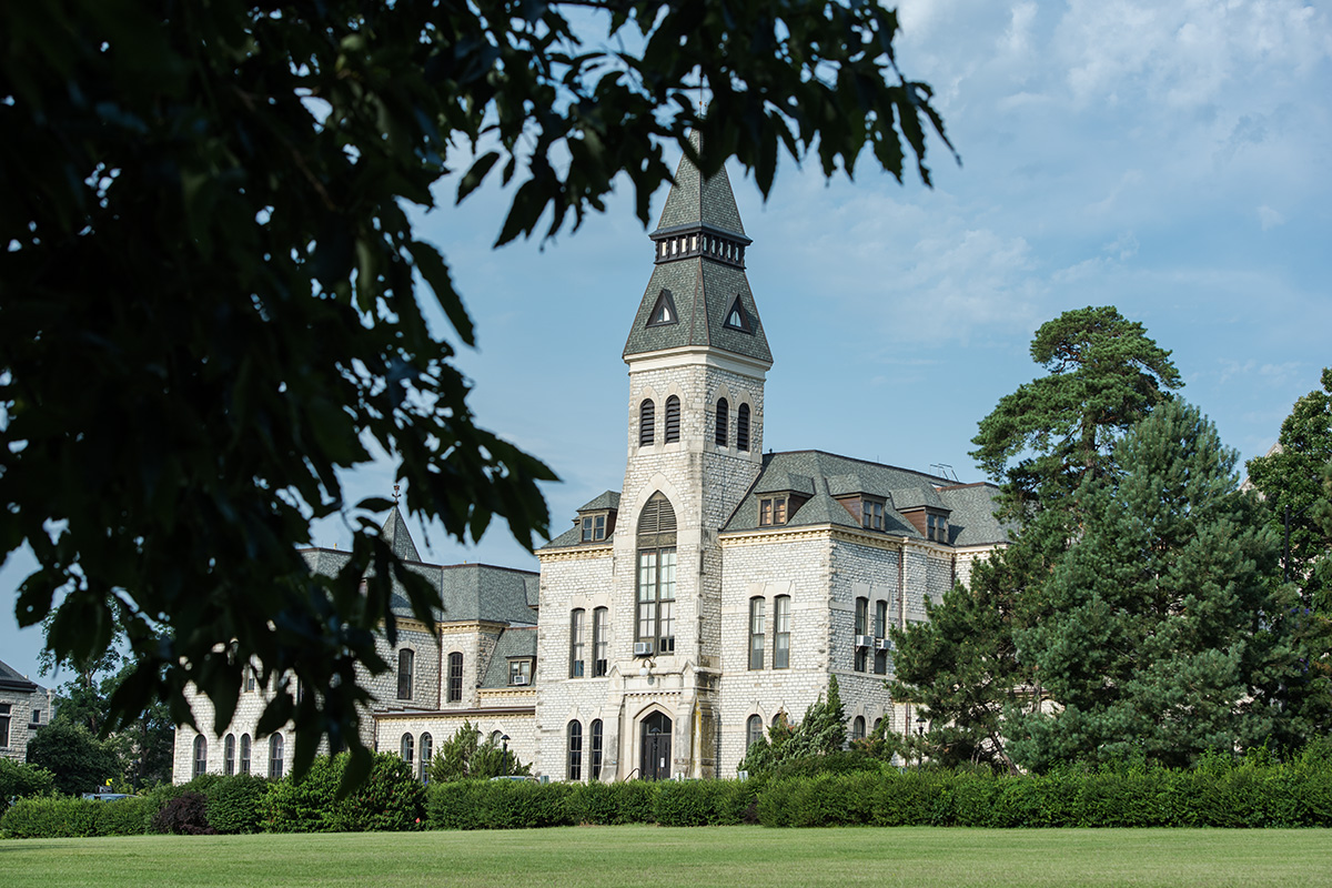 Looking over the lawn from behind the leaves of a tree on a sunny day at Anderson Hall at Kansas State University, a limestone building with a central clock tower.