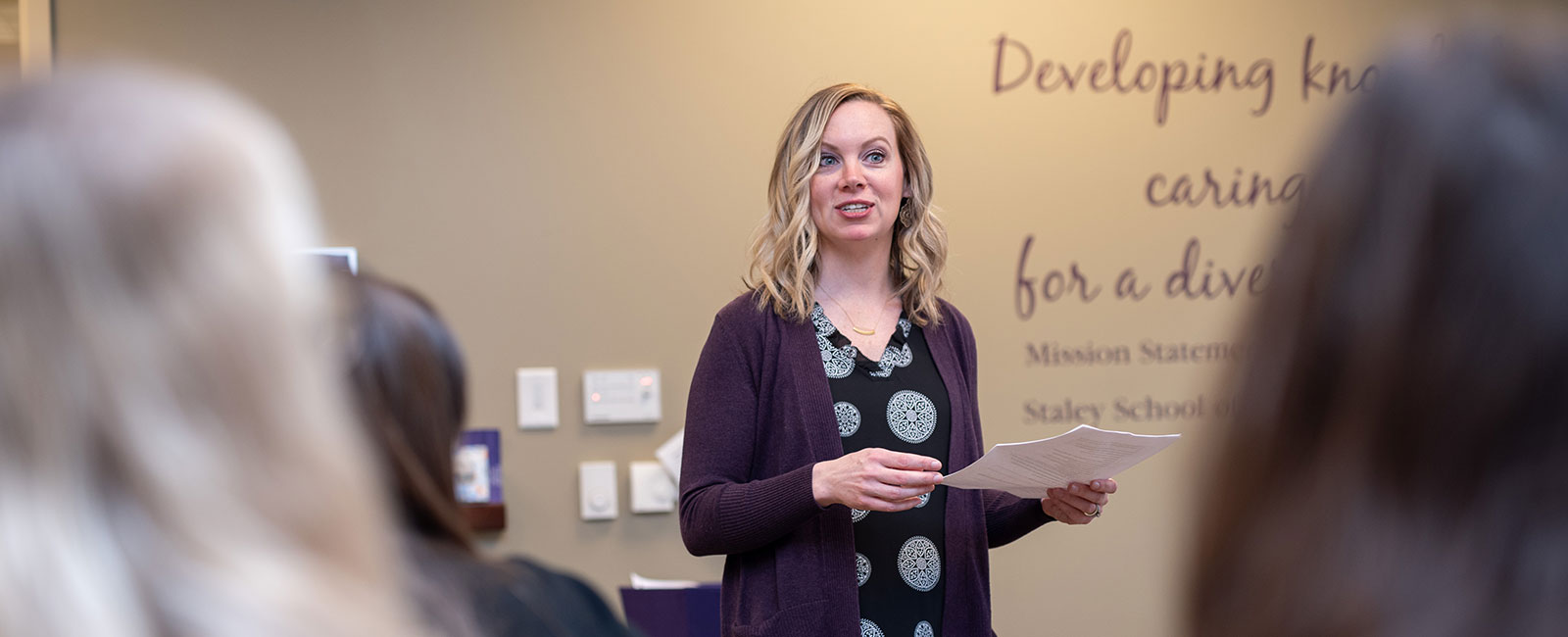 A woman in a black and white shirt with a purple cardigan speaks to a class.