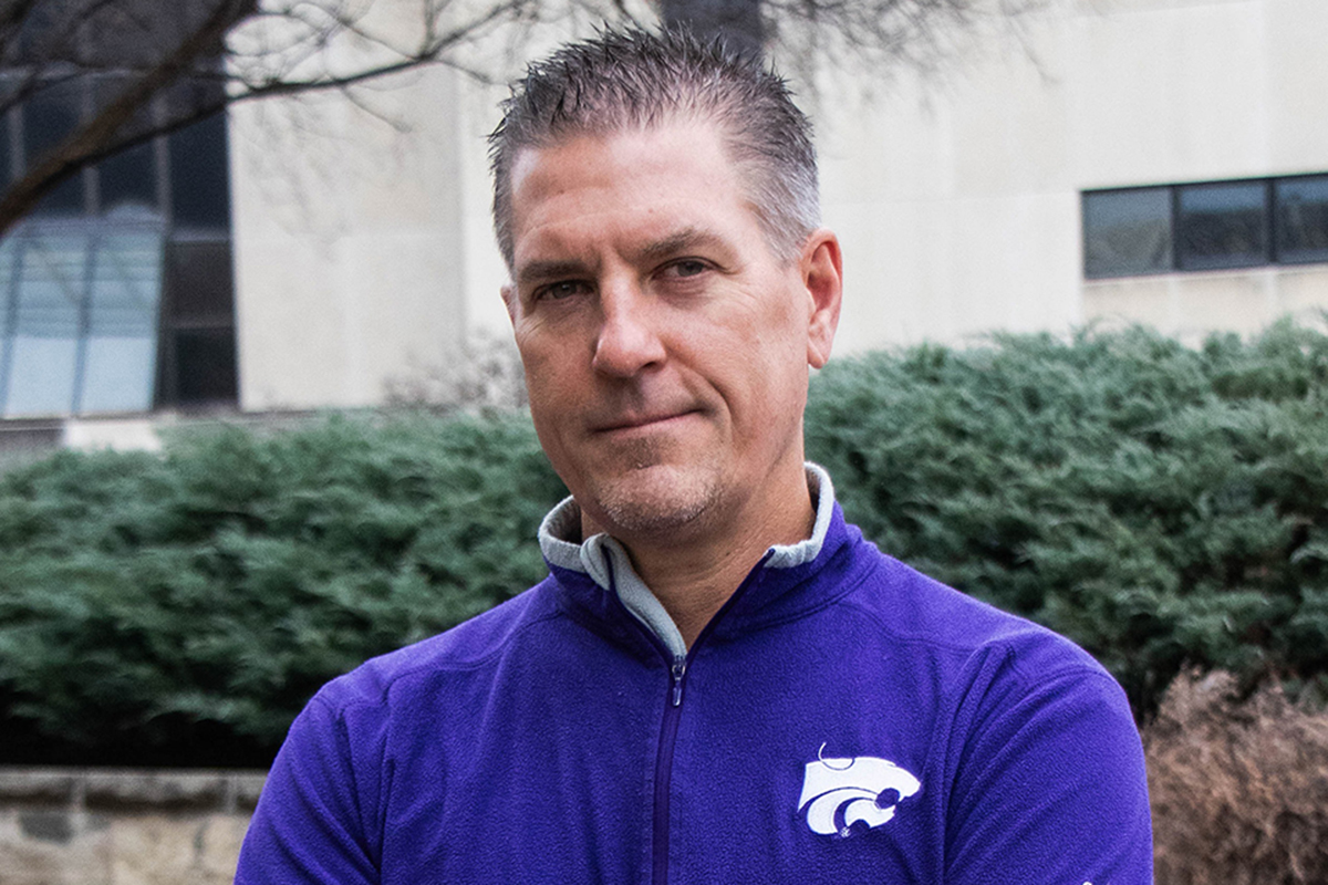A male college professor in a purple fleece jacket poses for an outdoor portrait.
