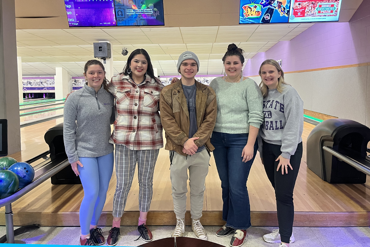 Five college students pose for a group photo in front of a bowling alley lane.