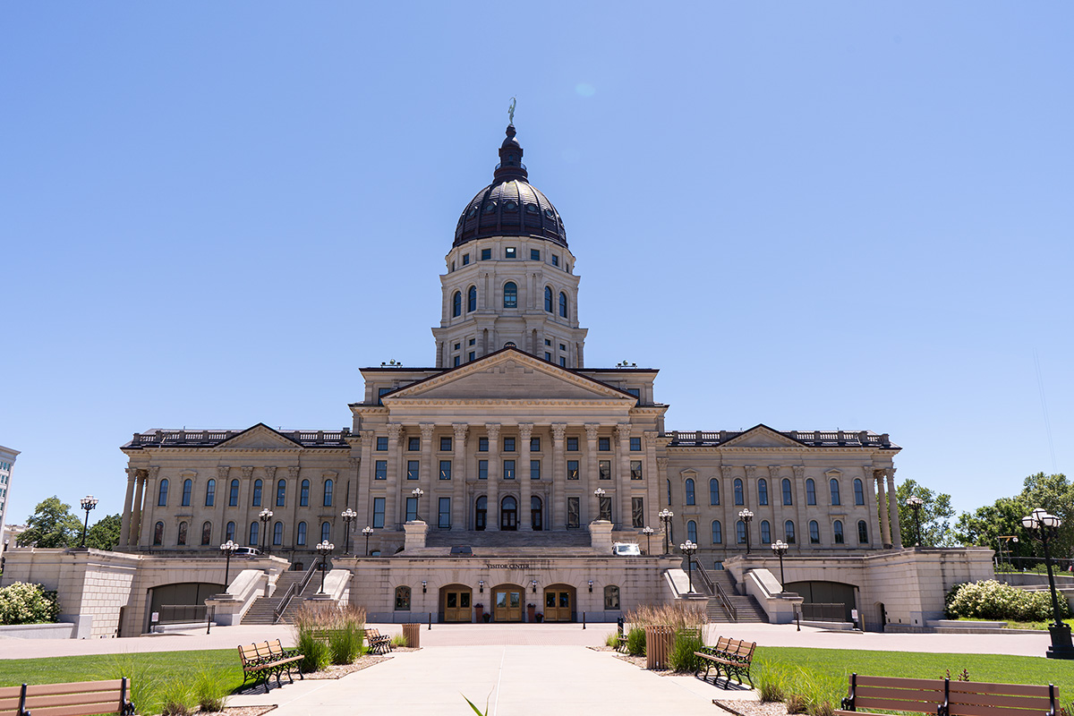 The Kansas Capitol, a rectangular building with a dome on top, stands against a blue sky with the walkway to the front door and green lawns shown in front.