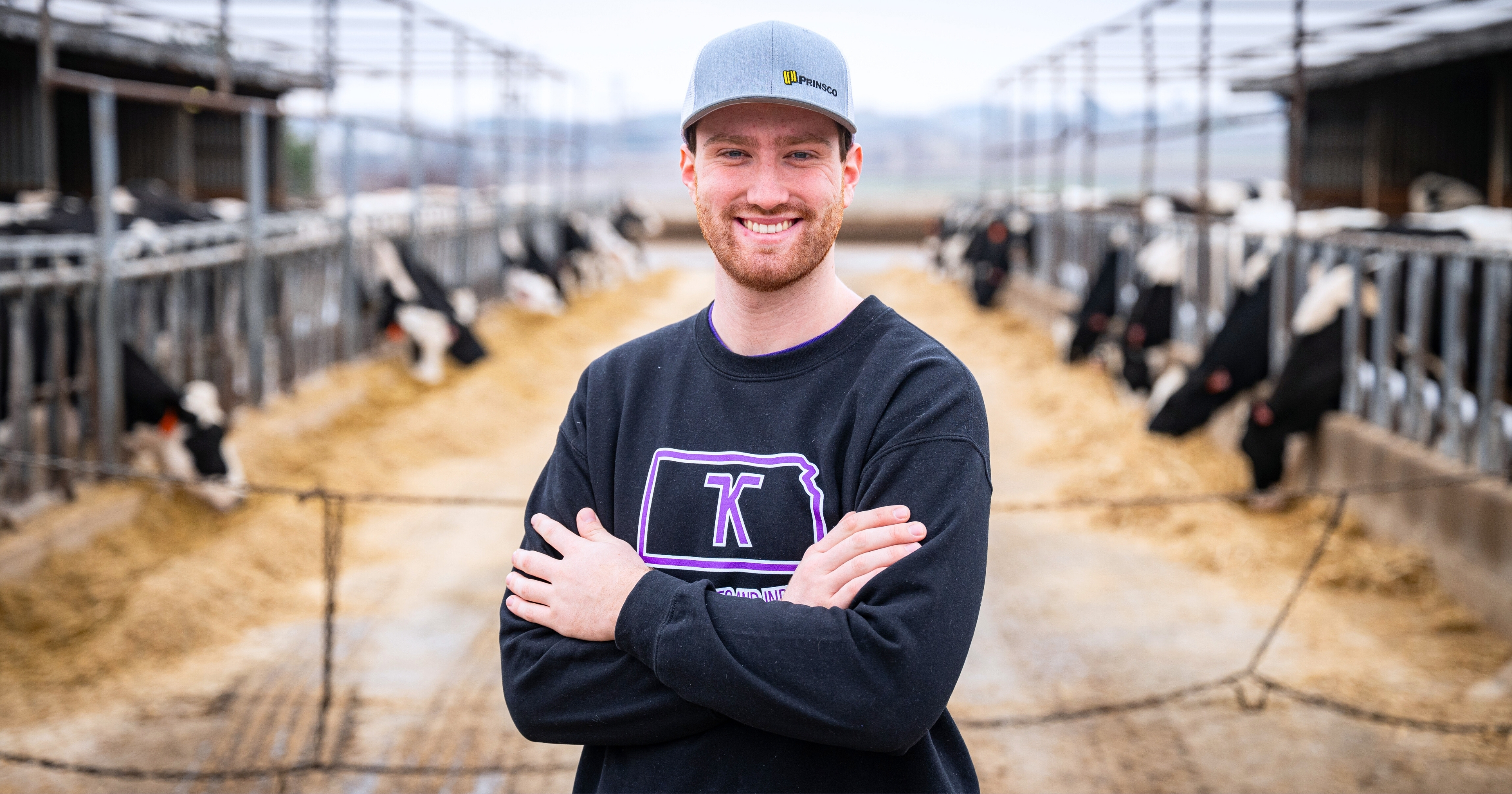 Carson Corwin, wearing a black sweatshirt and gray baseball cap, smiles with his arms crossed while standing in a feedlot aisle lined with Holstein cows in the background.