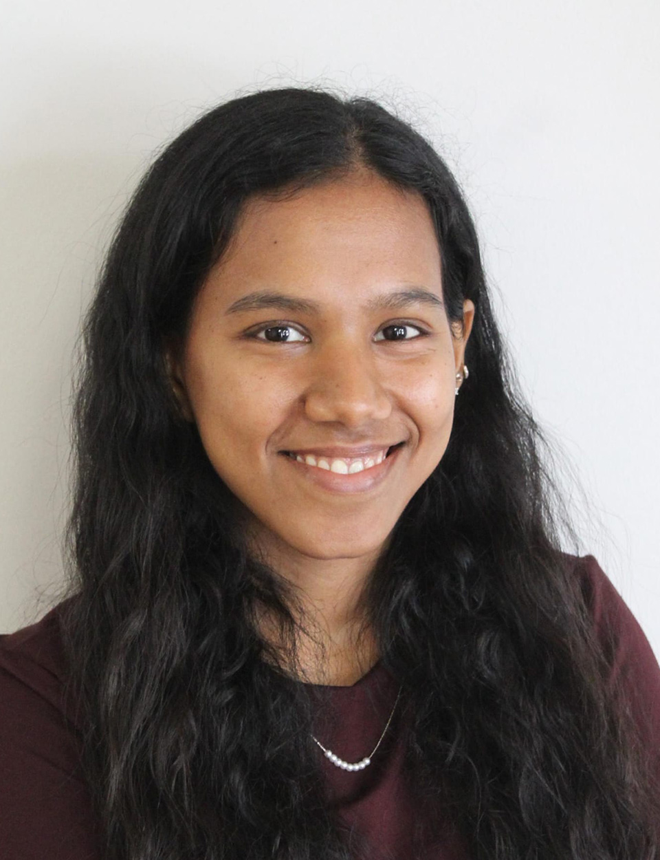 A graduate student with long black hair wearing a maroon shirt and necklace smiles against a white background.