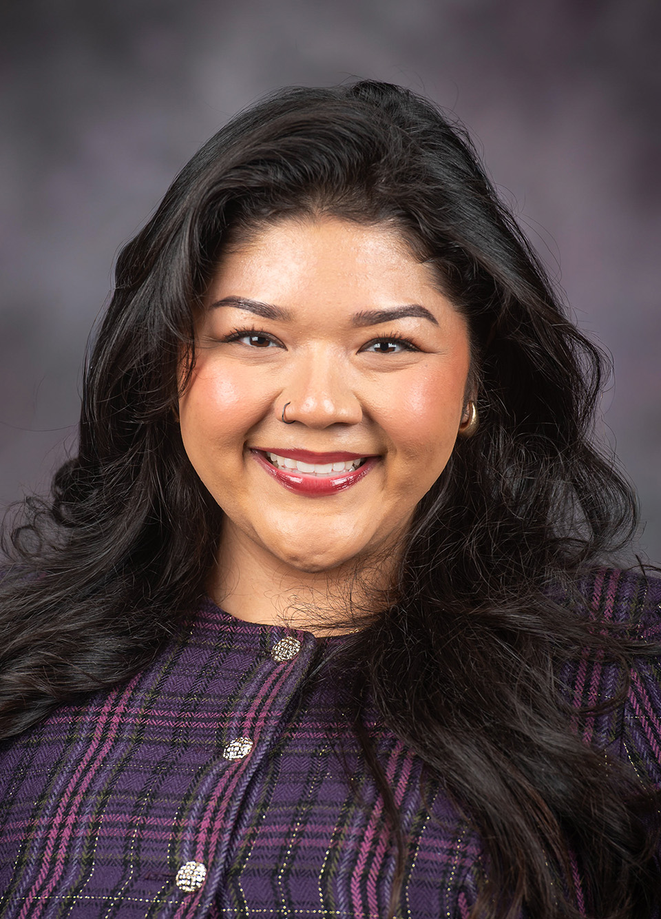 A graduate student with long black hair wearing a purple and pink plaid shirt smiles against a dark grey backdrop.