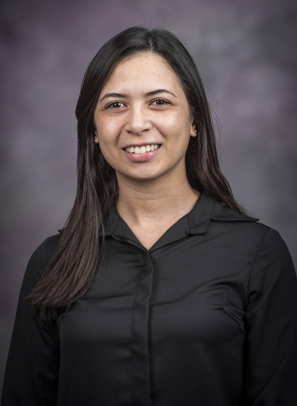 A graduate student with shoulder-length black hair wears a black collared shirt and smiles against a dark gray backdrop.