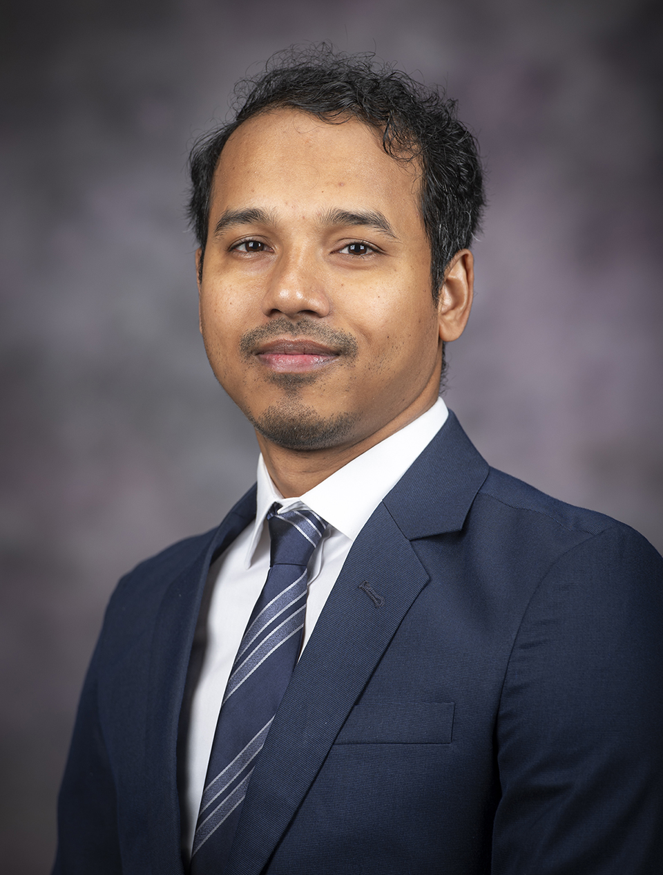 A graduate student with black hair wears a navy suit and tie against a grey backdrop.