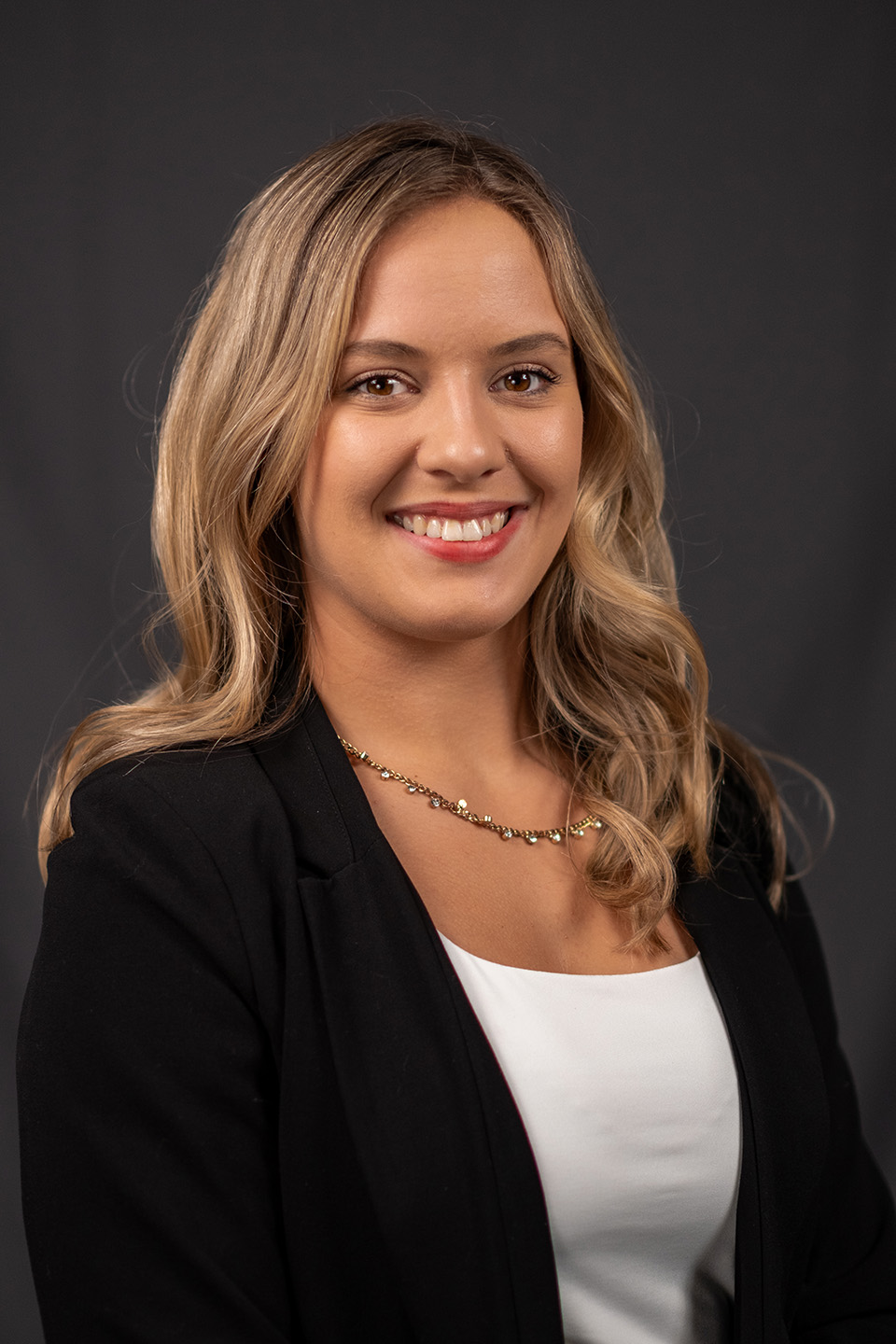A graduate student with long blonde hair wears a black blazer over a white shirt with a gold necklace and smiles against a dark backdrop.