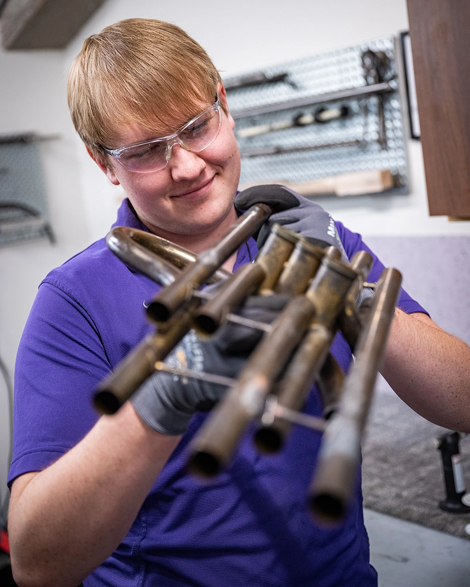 A male college student holds the curbed end of a musical horn up as he examines a brace he soddered onto it.