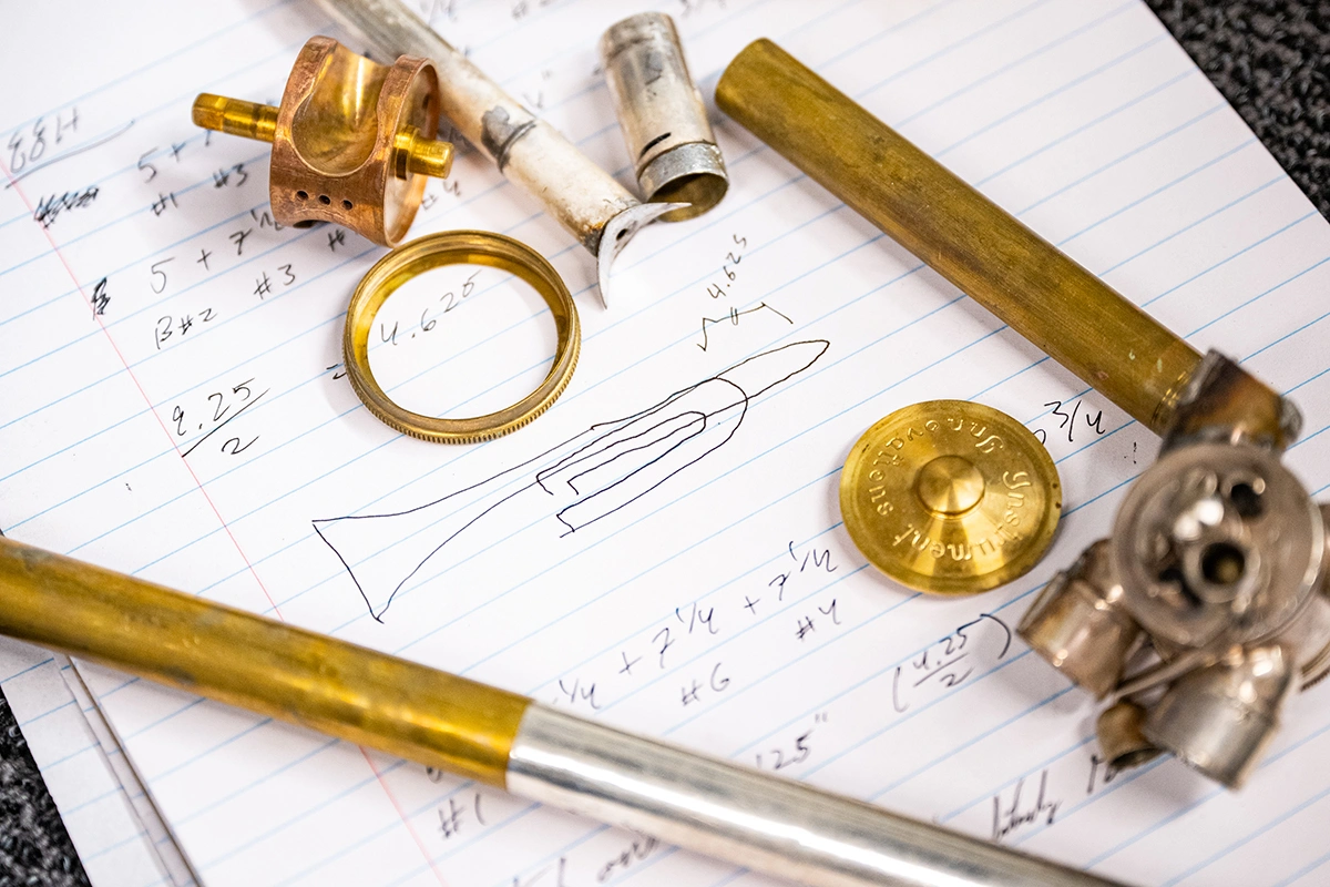 A top-down view shows several brass tubes and valves laying over a notepad that has various mathematical formulas written on it in pencil, as well as a basic sketch of the shape of a trombone.