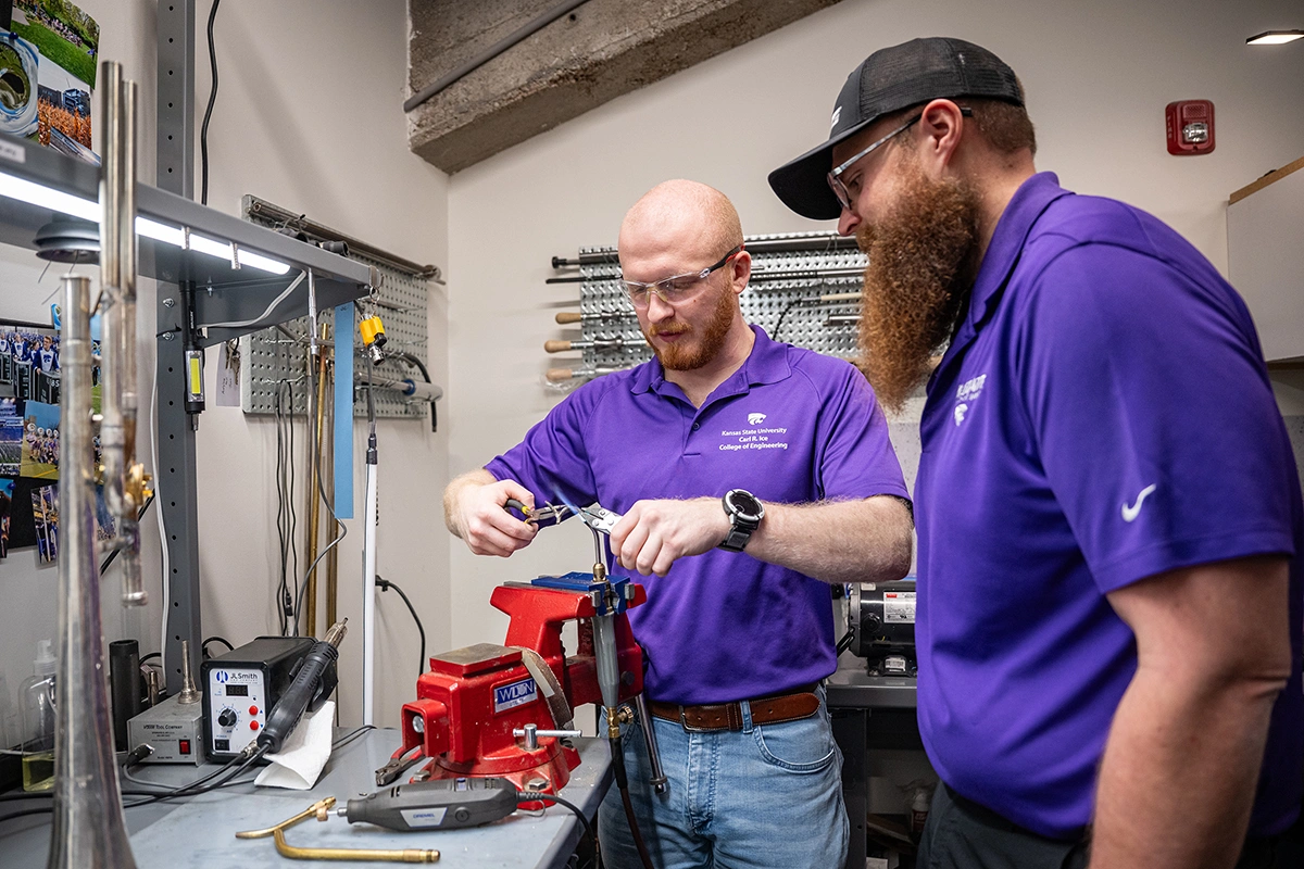 A bearded male college instructor checks the work of a student who is working on a musical instrument repair at a workbench in a repair workshop.