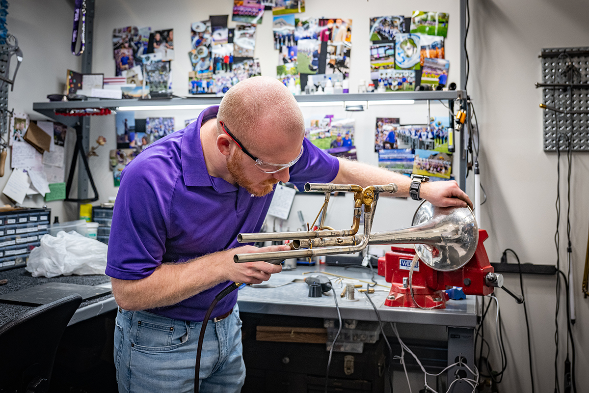A male college student in a purple polo uses a bench tool to balance and hold a trombone along its length as the student sodders a piece onto the instrument. On the wall behind the bench, several colorful photos show scenes from a marching band's season of performances.