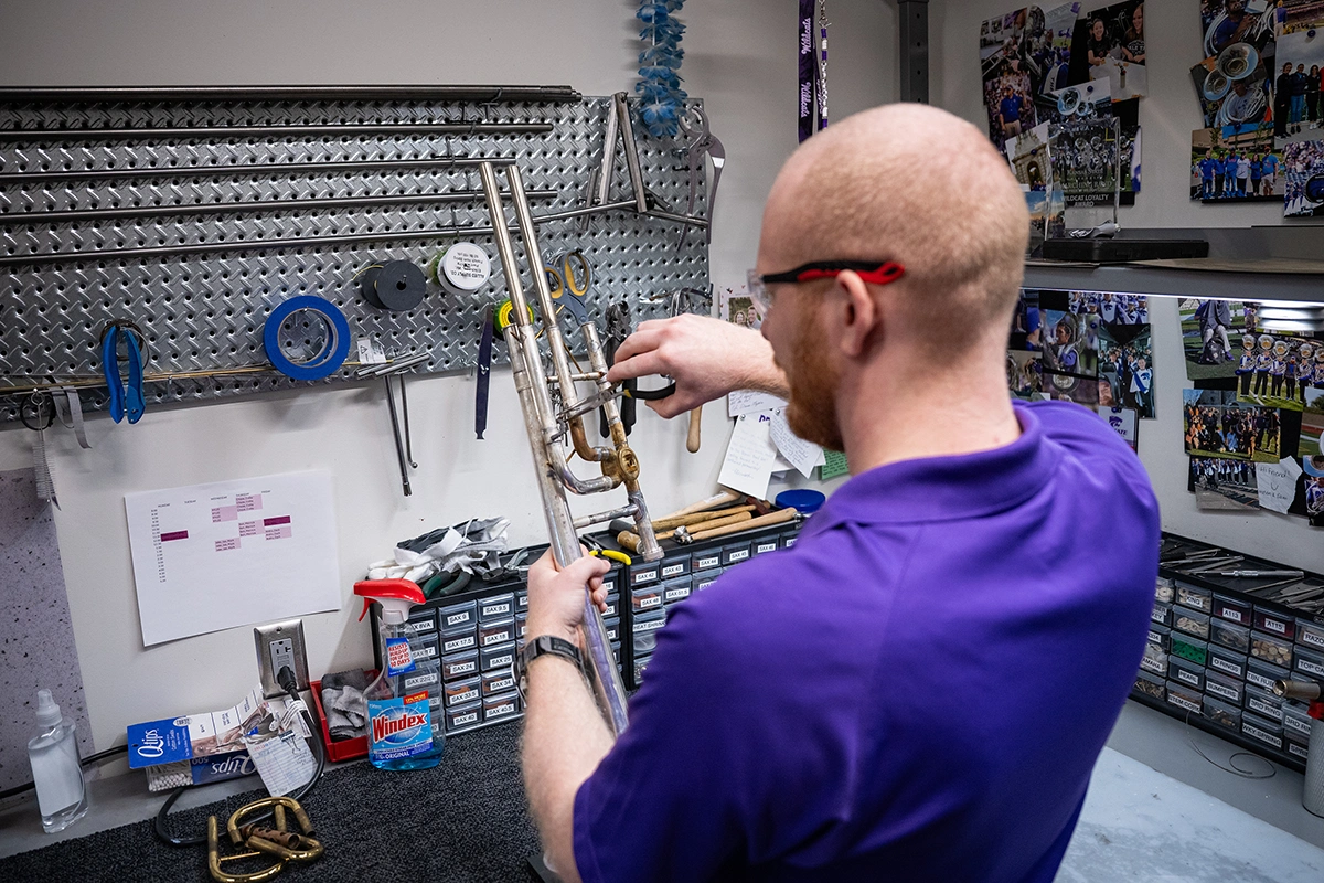 At a musical instrument repair workbench, a male college student in a purple polo and safety glasses uses a tool to measure the space between gaps in a metal horn.