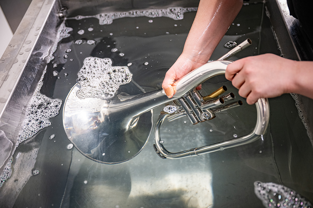 With a few bubbles on top, a melophone gleams underneath a solution of grayish blue liquid in steel tub while a female college student holds the instrument.