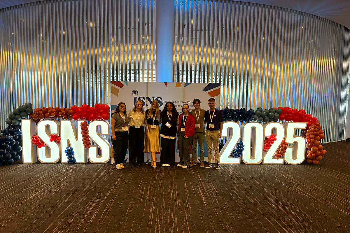 Seven college students stand in front of a large photo backdrop that says "ISNS 2025" in illuminated letters. 