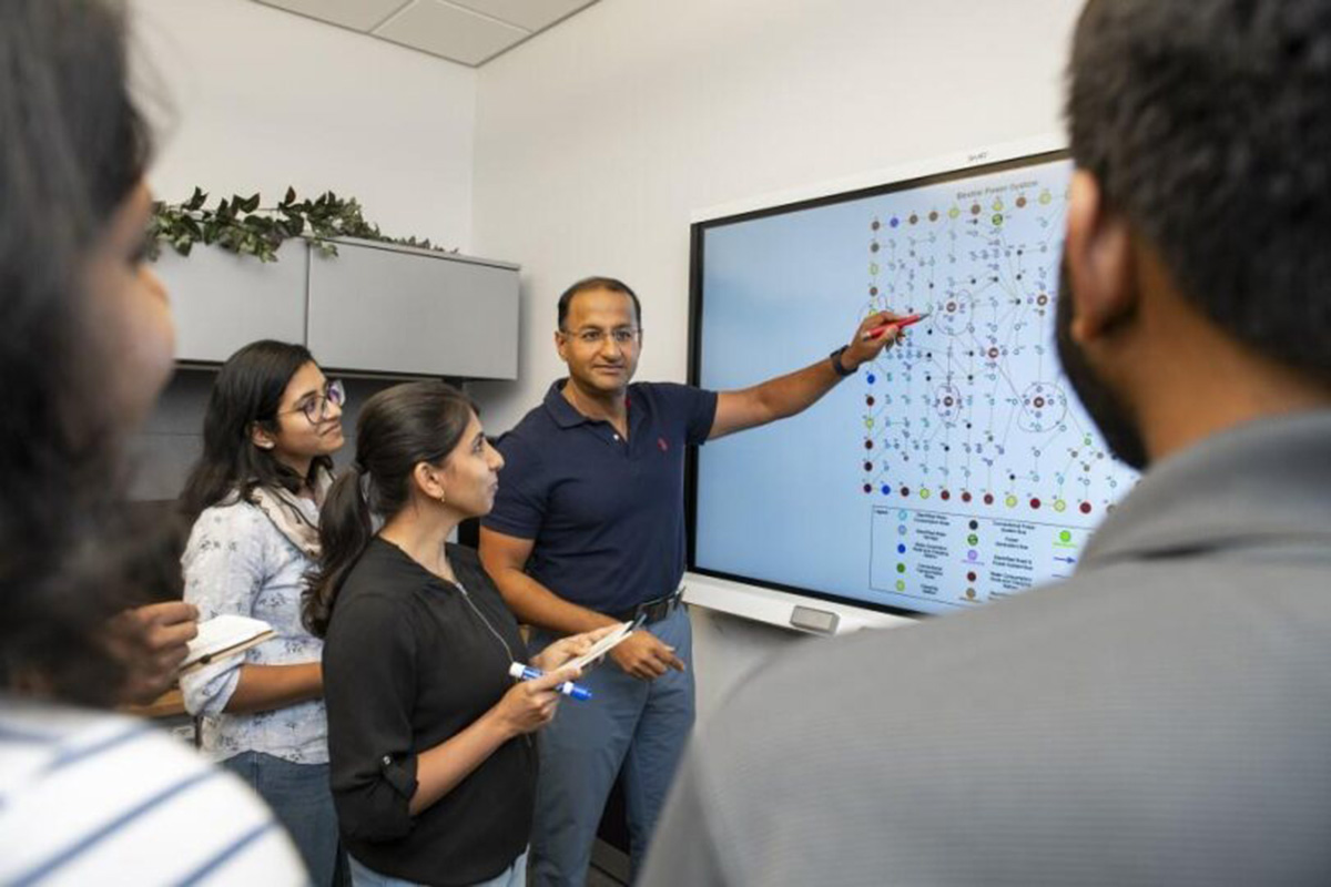 A researcher stands in front of a whiteboard and talks to a group of people in a classroom. 