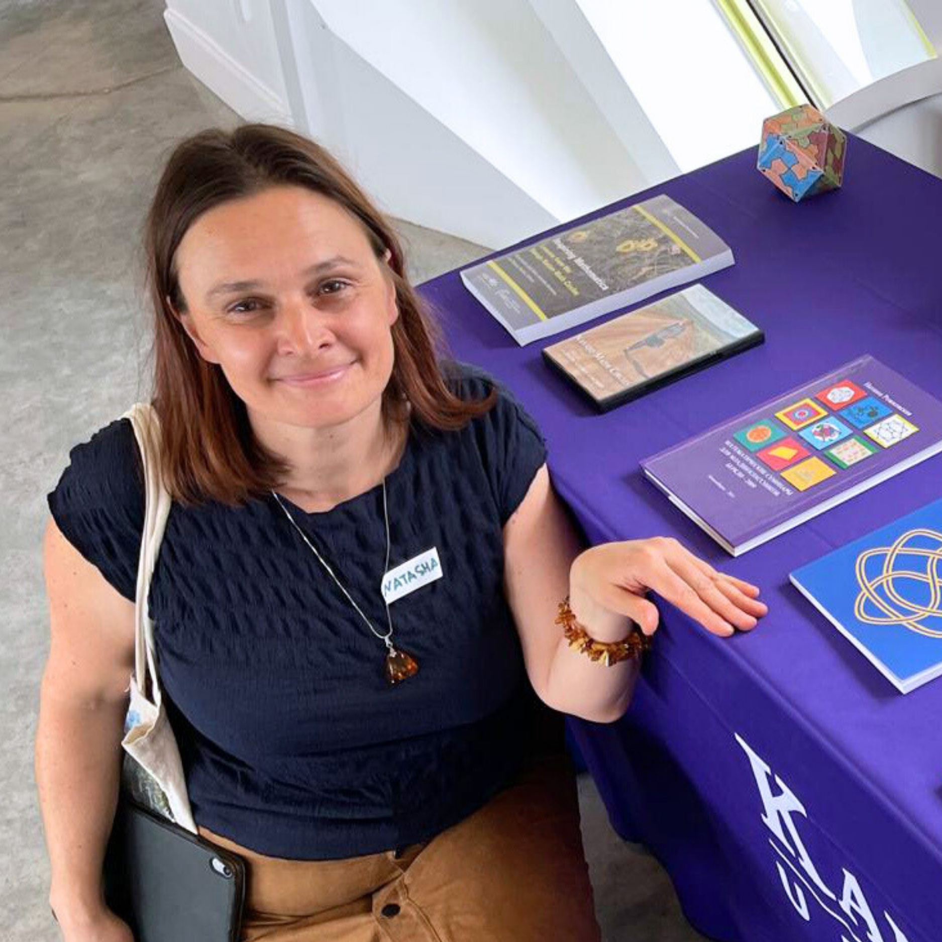 Natasha Rozhkovkaya, wearing a navy blouse, crouches next to a table displaying her authored books during a Beach Museum of Art event.