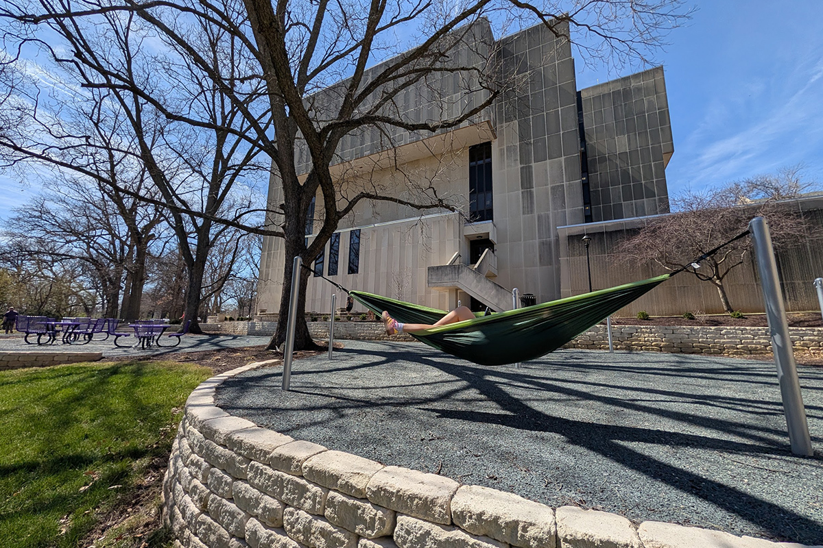 A college student lies in a green hammock in a new, graveled study area underneath a leafless tree by a large concrete academic hall on a college campus.