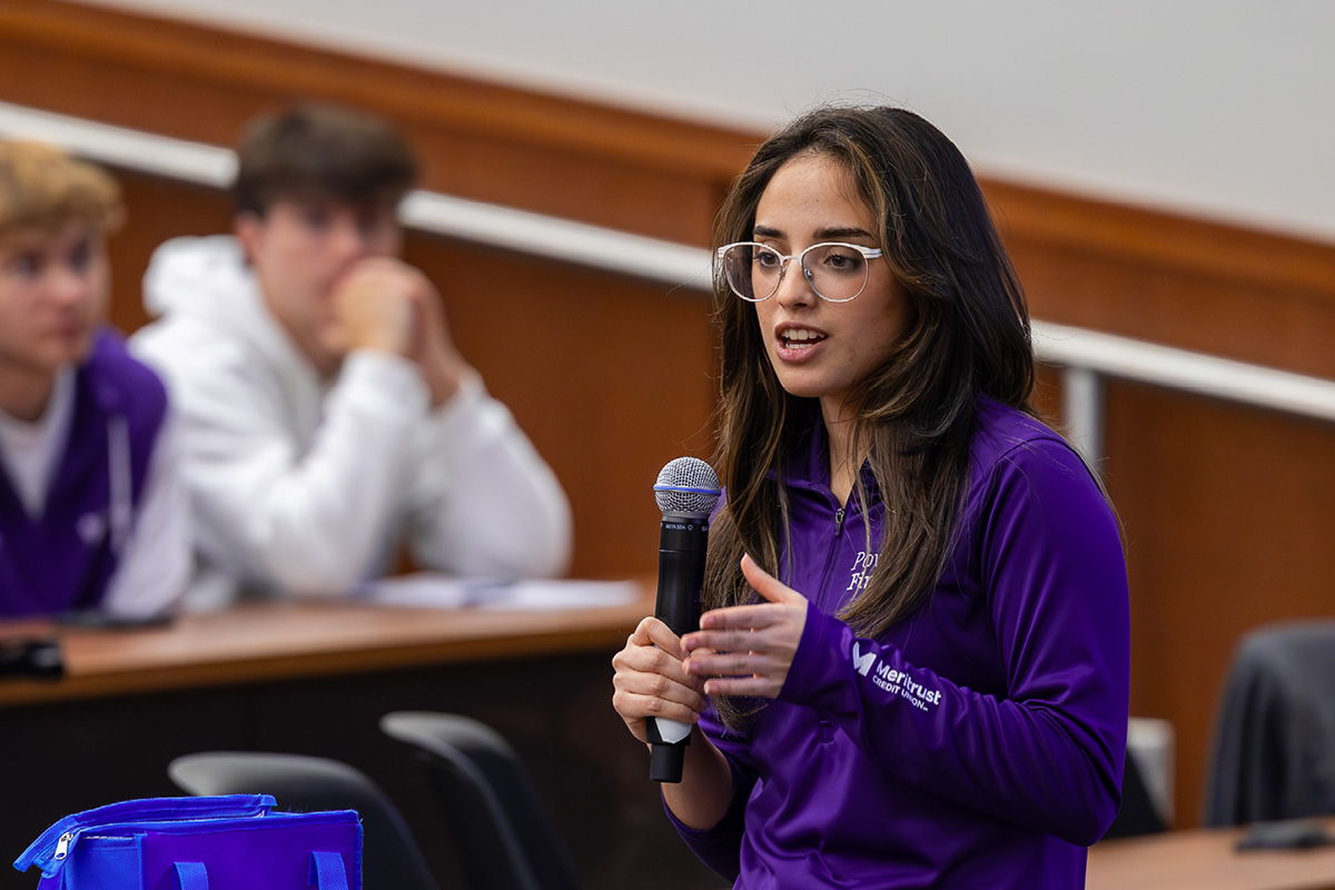 A female college student stands in front of a lecture hall and addresses other college students. She has brown hair, is wearing glasses and a purple ¾-zip jacket, and is holding a microphone.