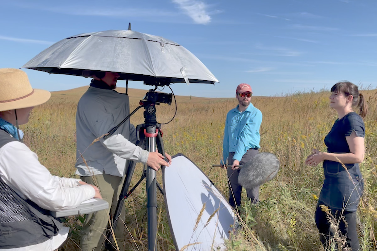 A film crew records an interview in a tallgrass prairie, with a camera on a tripod, boom microphone and reflector visible as a woman speaks on the right under a clear blue sky.