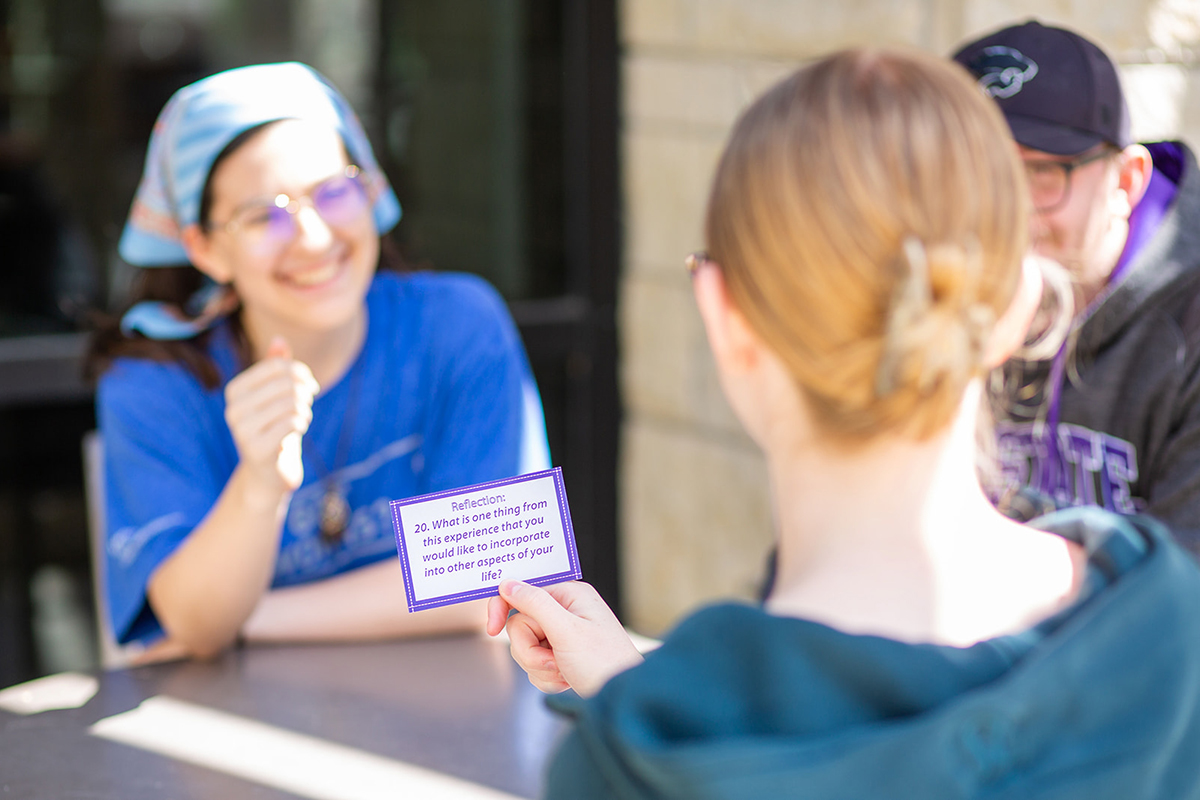 A college student holds a conversation starter card as she facilitates an outdoor community building activity.