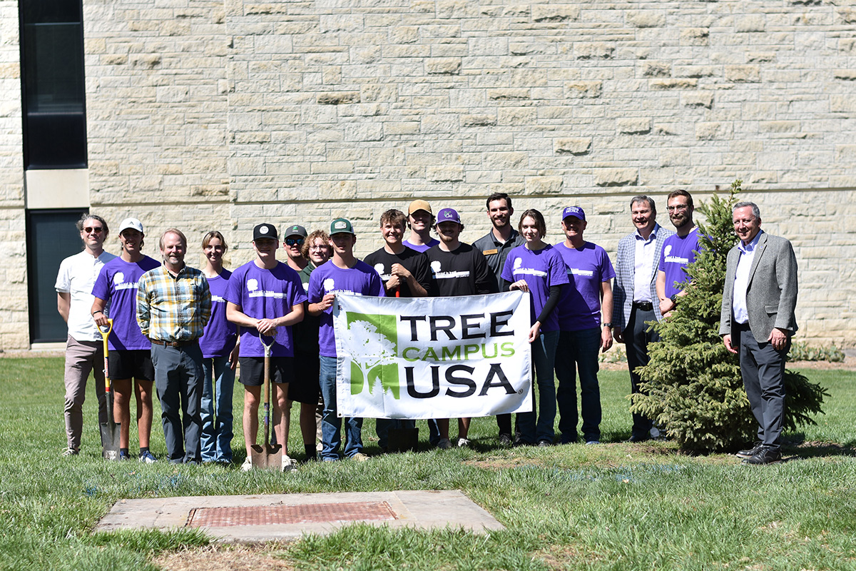 A group of college students and university employees stand outside a limestone building on a sunny day with a white banner that reads 
