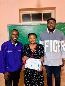 Three people stand together in a classroom and the woman in the middle holds a certifcate. 