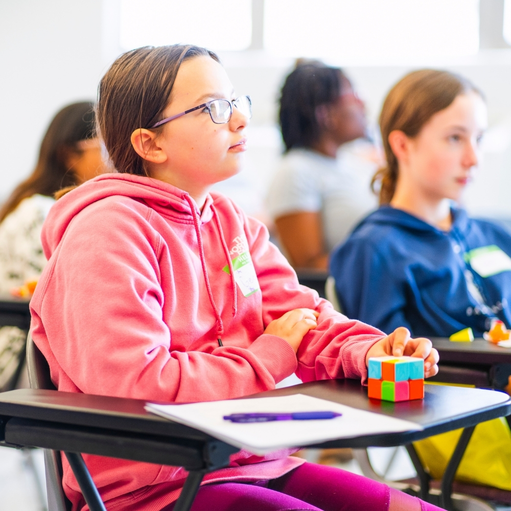 A middle school student in a pink hoodie and glasses sits at a desk, looking attentively toward the front of a bright classroom while holding a colorful cube puzzle. Other students, slightly out of focus, are in the background.