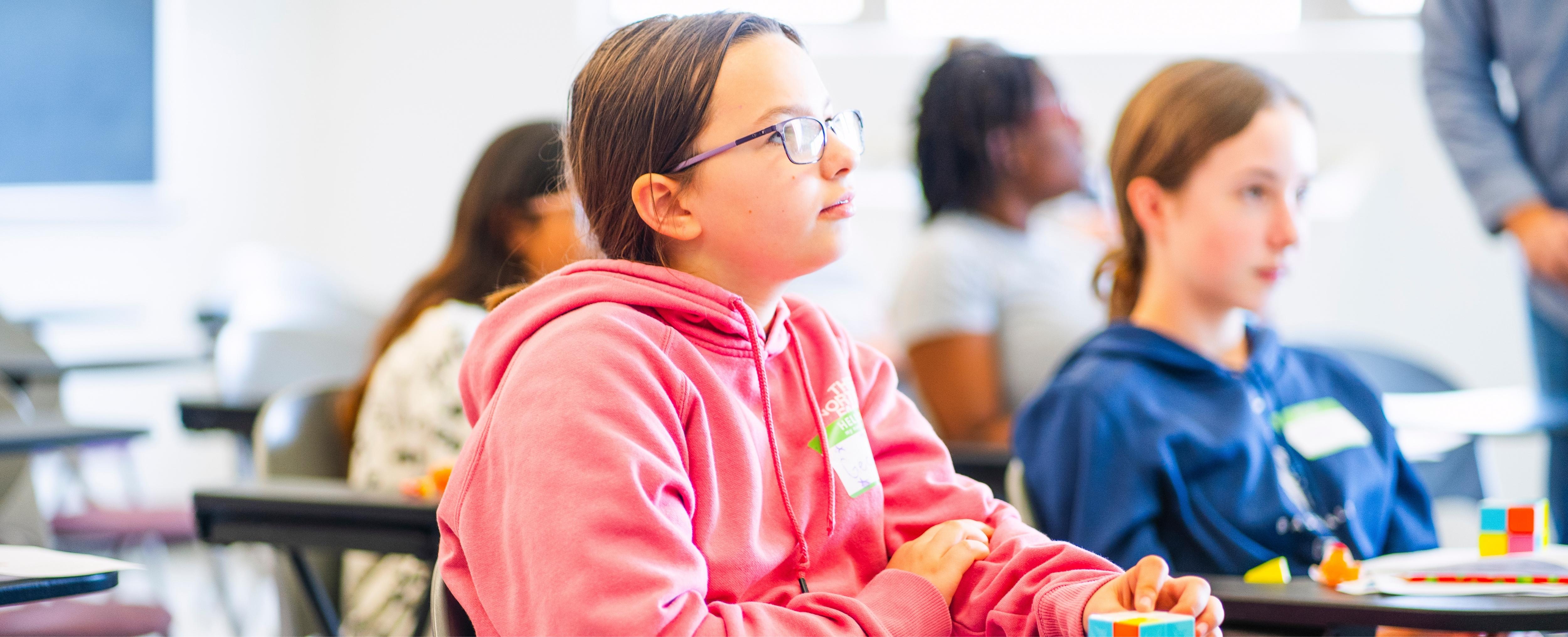 A middle school student in a pink hoodie and glasses sits at a desk, looking attentively toward the front of a bright classroom while holding a colorful cube puzzle. Other students, slightly out of focus, are in the background.