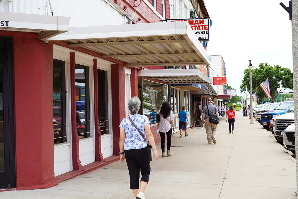 People are shows walking down a rural business block that is lined by parked cars and American flags hanging from ligthpoles.