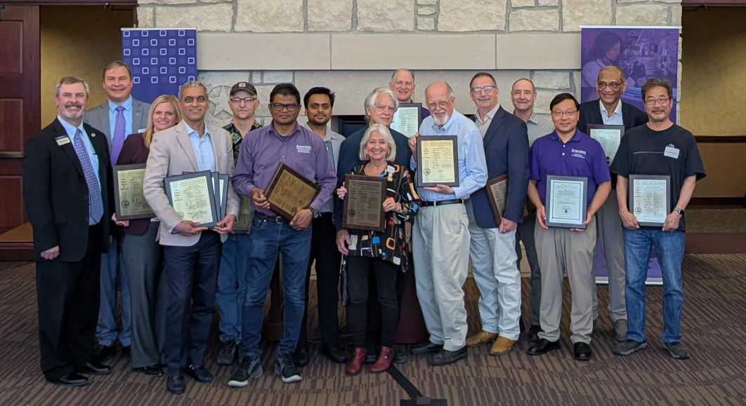 Group photo of 15 people standing indoors in front of a stone fireplace, many holding framed certificates and plaques during an awards ceremony. Purple Kansas State University banners are on either side of the group.