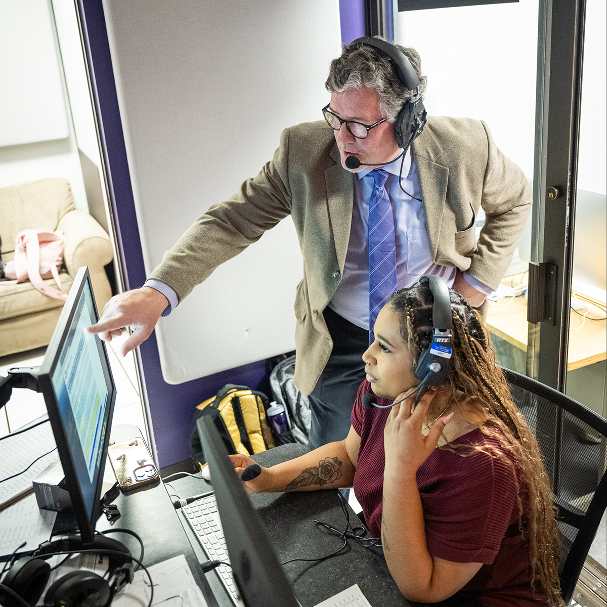 A male college professor assists a female college student who is managing a control panel in a television production studio.