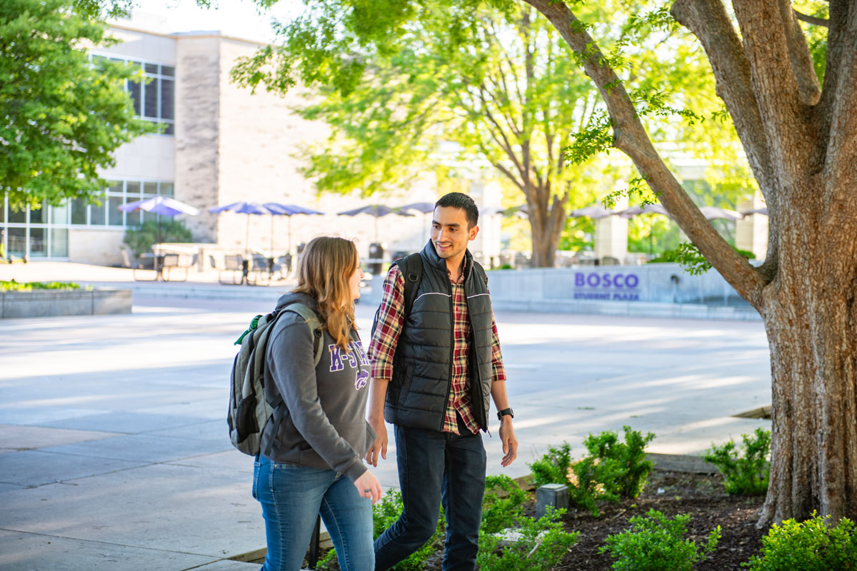 Two students walk together in Bosco Student Plaza. One student is wearing a grey K-State sweatshirt and the other student is wearing a plaid shirt with a grey vest.