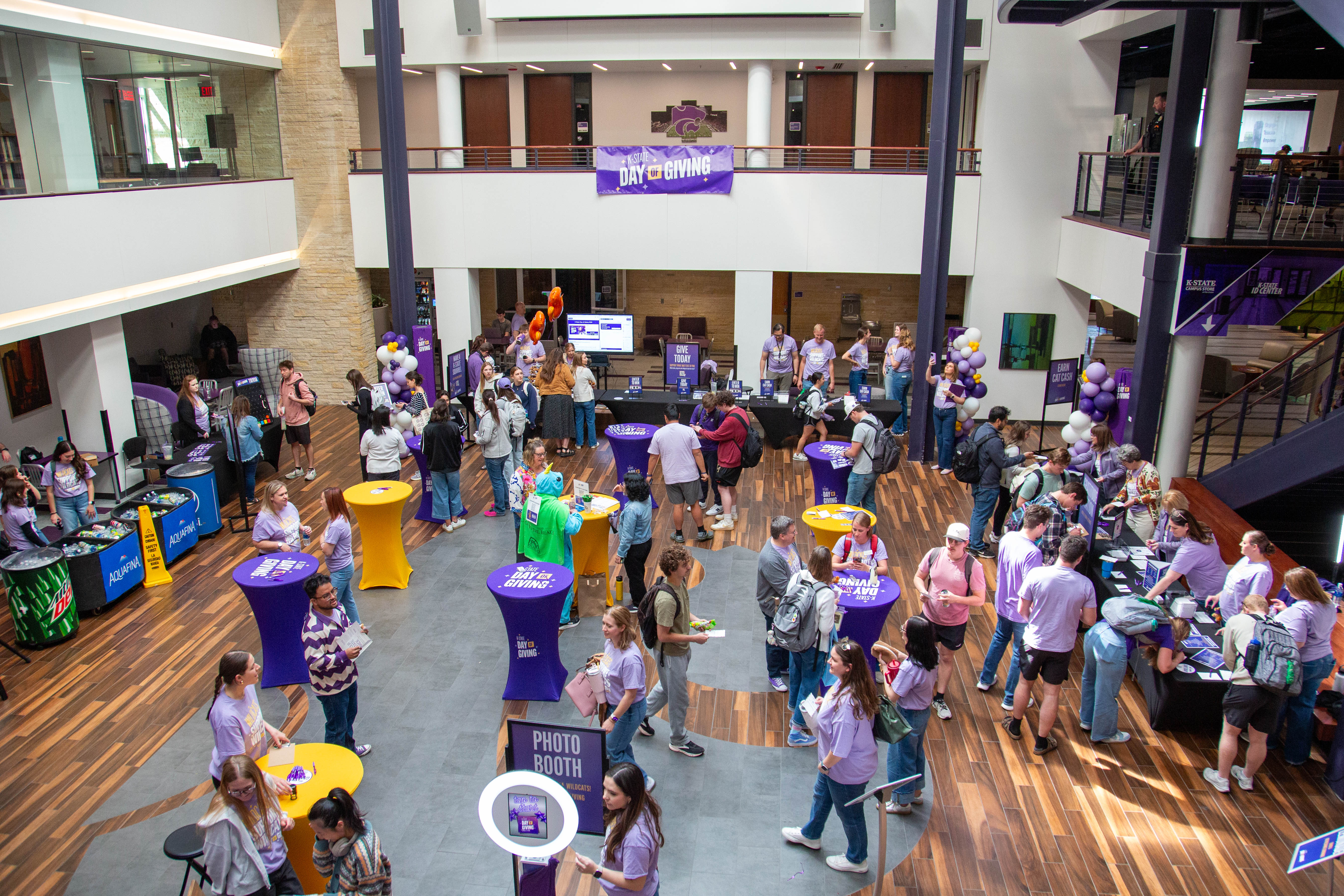 A downward view of the courtyard in the K-State Student Union shows people visiting booths and tables set up for K-State Day of Giving. Many people are wearing the themed lavender shirts.