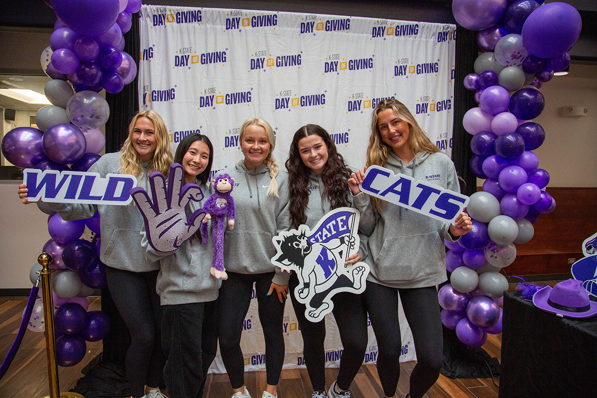 Five female students pose in front of a K-State Day of Giving backdrop for a photo booth. They are all wearing gray hoodies and holding up various purple props.