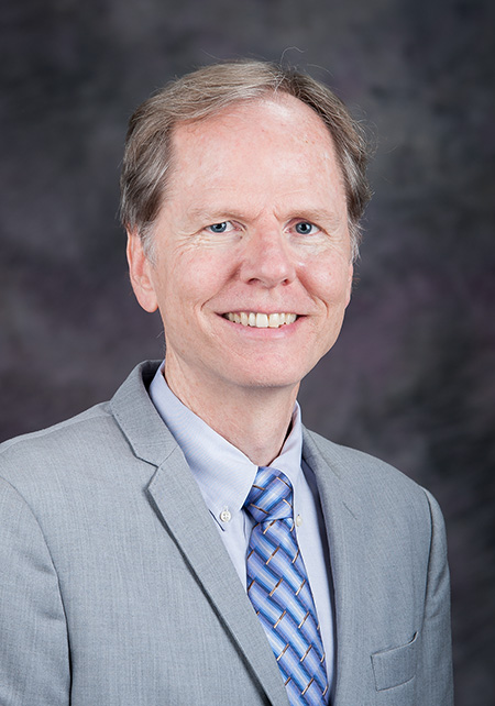 A man in a gray suit smiles against a dark gray backdrop.