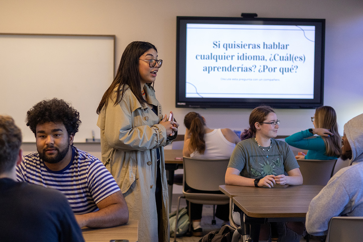 A graduate teaching assistant wearing a tan long coat walks between students sitting at desks and teaches a Spanish lesson.