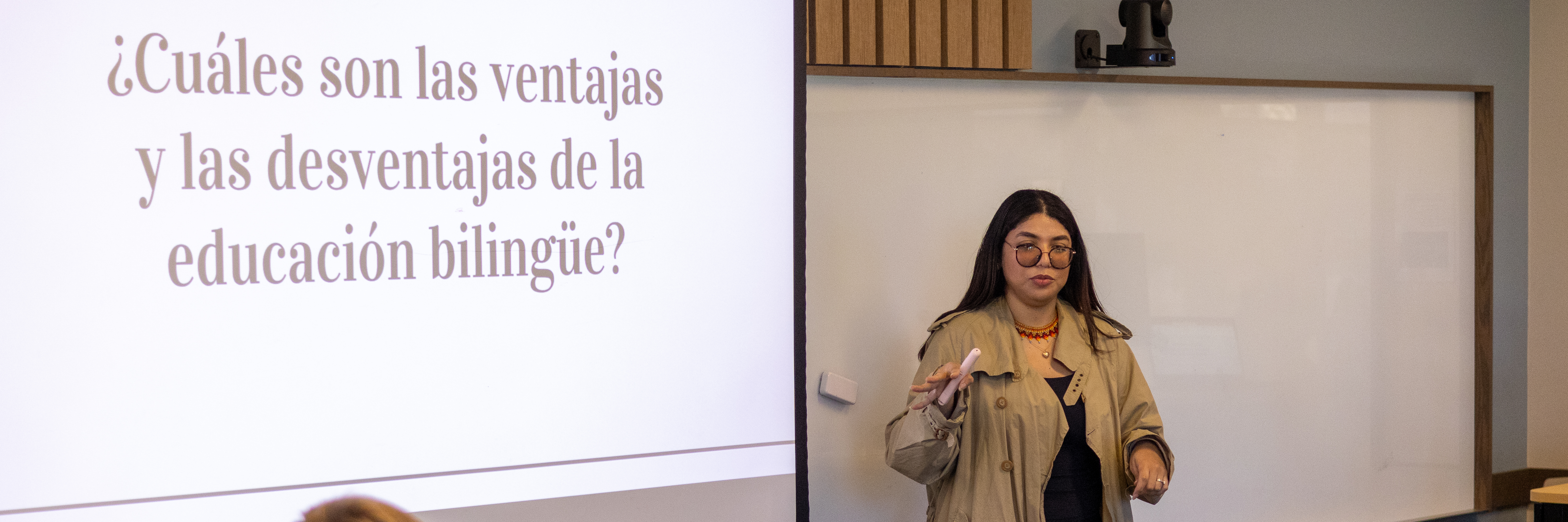 A graduate teaching assistant wearing a tan long coat walks between students sitting at desks and teaches a Spanish lesson.