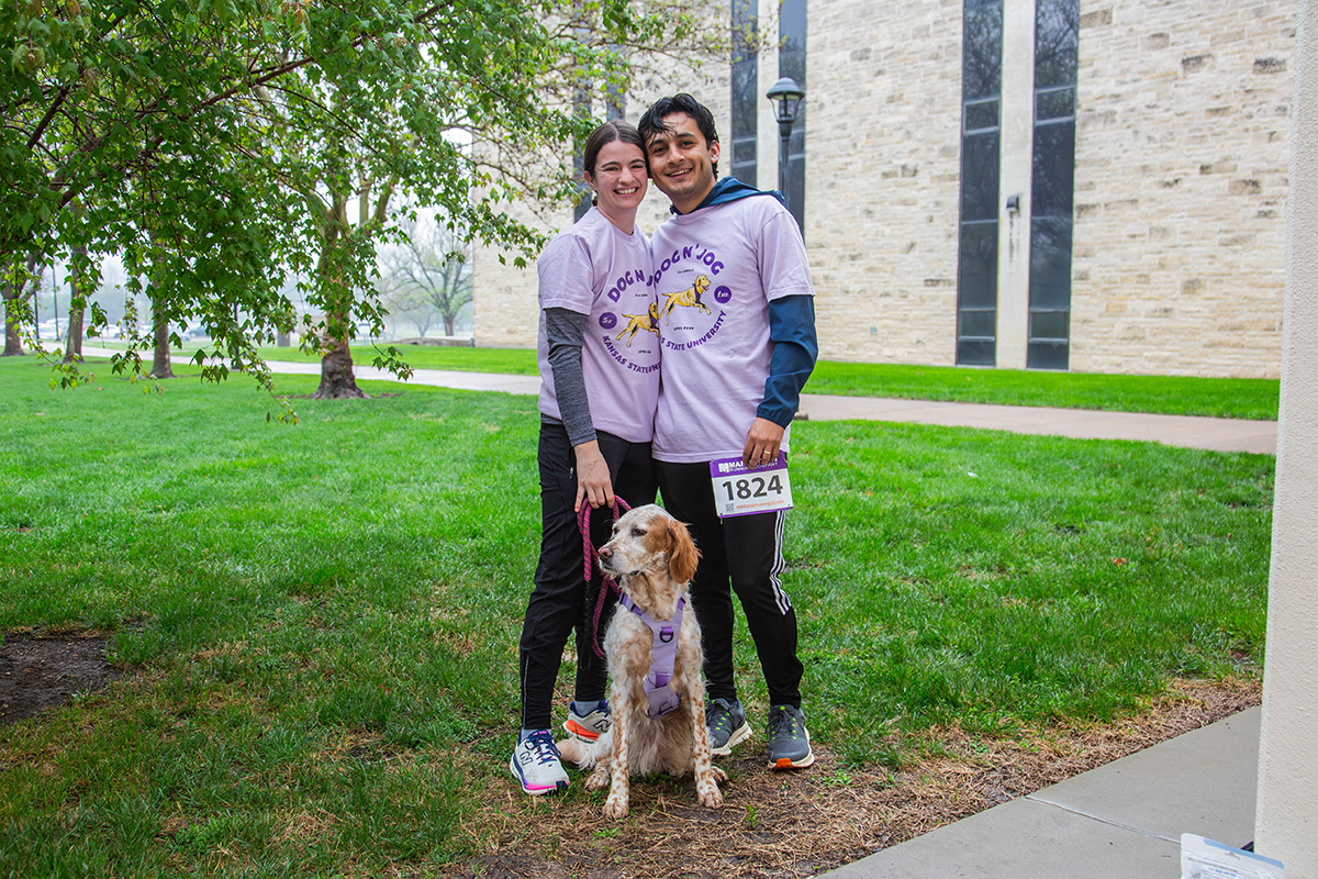Two smiling participants wearing matching purple "Dog-N-Jog" t-shirts pose with their brown and white spaniel wearing a purple harness on green lawn. Race bib number 1824 visible on runner's leg.
