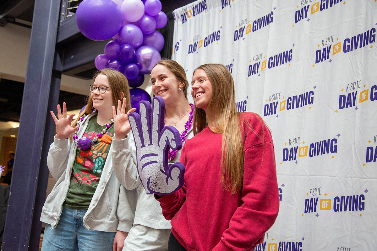 Three smiling students pose in front of a white "K-State Day of Giving" step-and-repeat banner, with two making the Powercat hand gesture and one holding a large foam Powercat hand prop. Purple balloon decorations are visible on the left side.