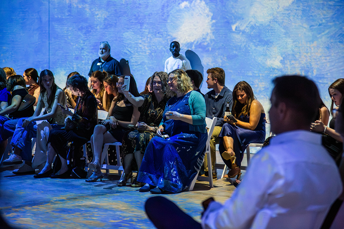 Audience members seated on white folding chairs watch fashion show presentation. Large projected backdrop displays blue sky with white clouds creating immersive atmosphere. Attendees wear mix of formal and casual attire including black dresses, sequined garments, turquoise top, yellow dress, and navy clothing. 