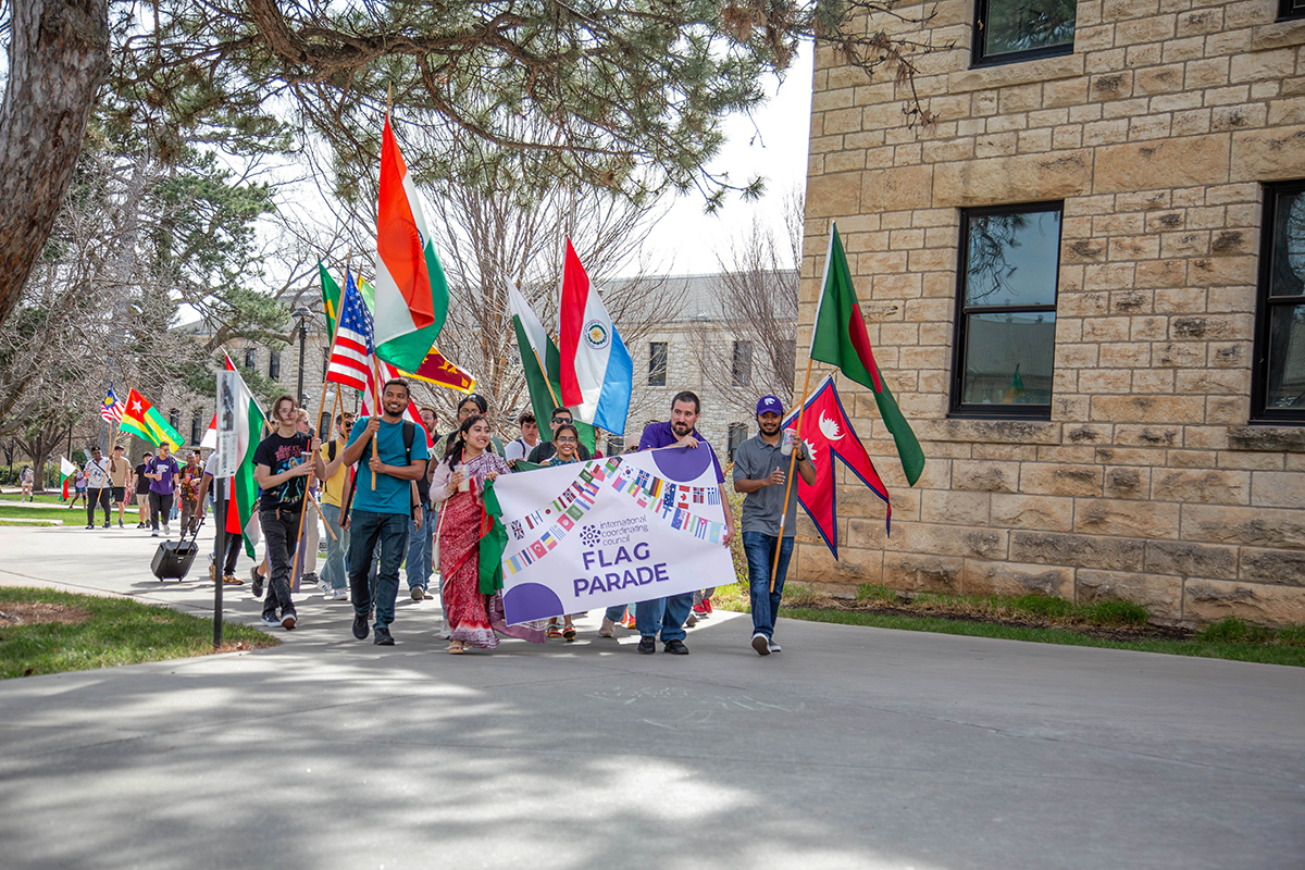 Group of students and staff march down campus sidewalk carrying international flags and white banner reading "International Student Week FLAG PARADE" with colorful flag graphics. Visible flags include United States, India, Nepal, and others held on wooden poles.