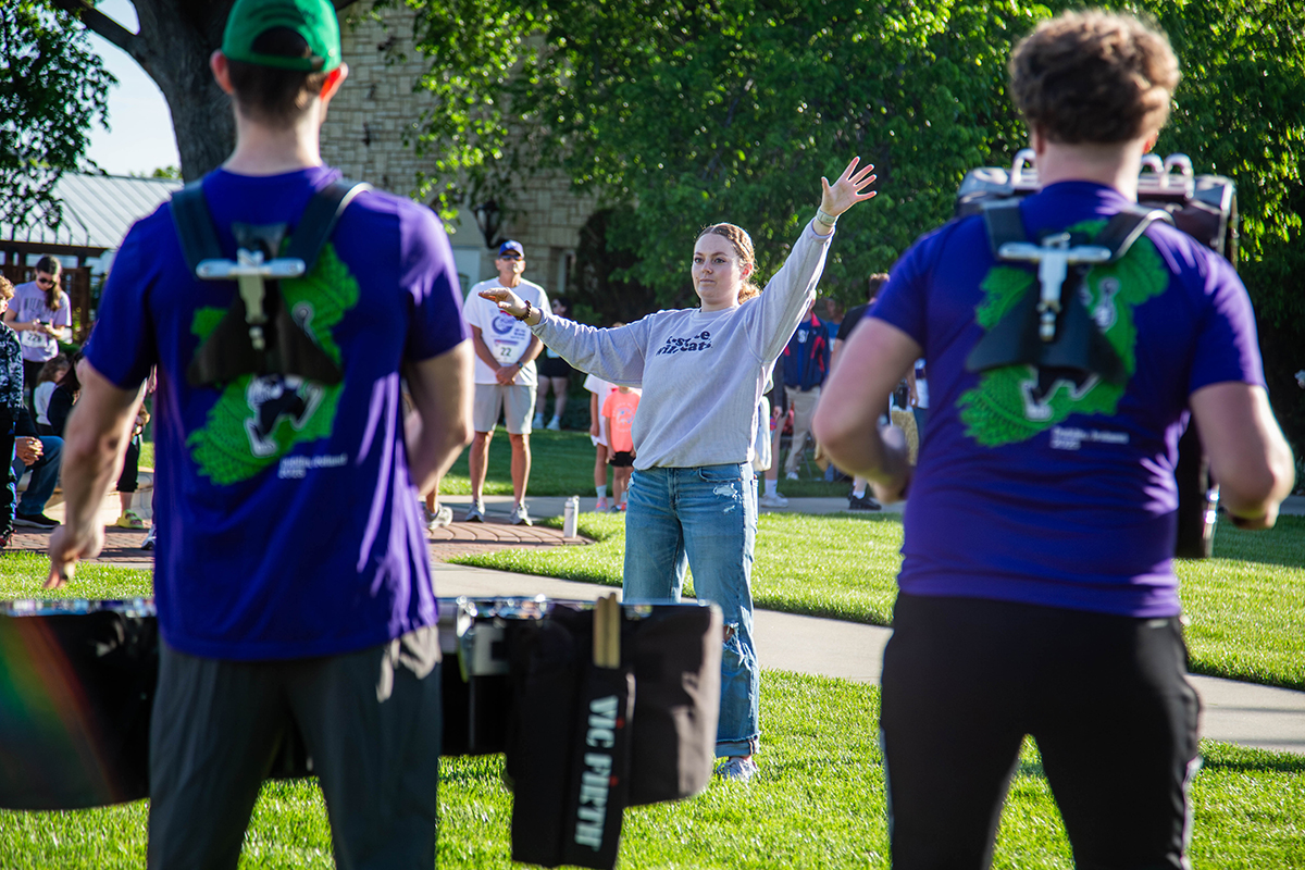 A drum major directs a line of drummers outdoors on a sunny, spring morning at a college campus.