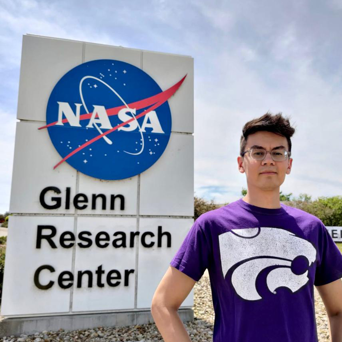 A male rocket engineer in a purple shirt with a K‑State Powercat symbol poses for a portrait in front of a bright blue NASA globe logo.